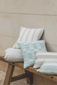 A collection of neutral and soft aqua-toned woven cushions arranged on a rustic wooden bench against a light stone wall, showcasing textured Perennials® performance fabrics in stripe and check patterns.